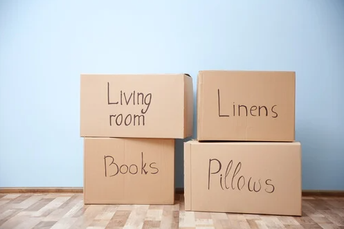 Four labeled moving boxes stacked against a wall reading Living Room, Linens, Books, and Pillows